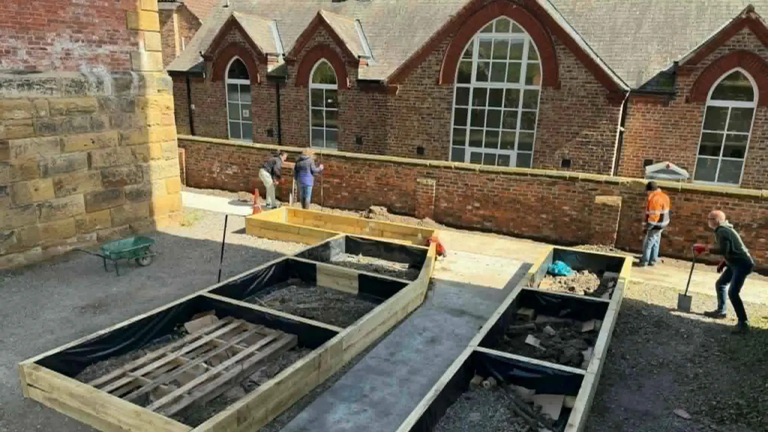 Supplied Four men and women using gardening tools to create a raised wooden flower beds area. There is an old stone building in the background, a brick building with four large windows and a brick wall.