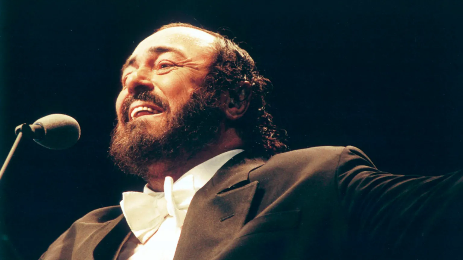  Pavarotti, dressed in distinctive opera jacket and white bow tie, singing into a microphone