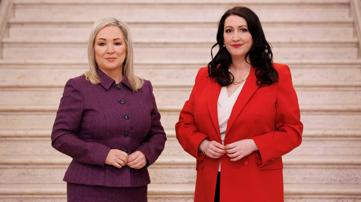  Michelle ONeill - who has shoulder-length blonde hair- in a purple suit and Emma Little Pengelly - who has longer dark brown hair - in a red suit with a white blouse visible under her jacket. They are standing in front of a set of marble stairs in the Great Hall at Stormont.