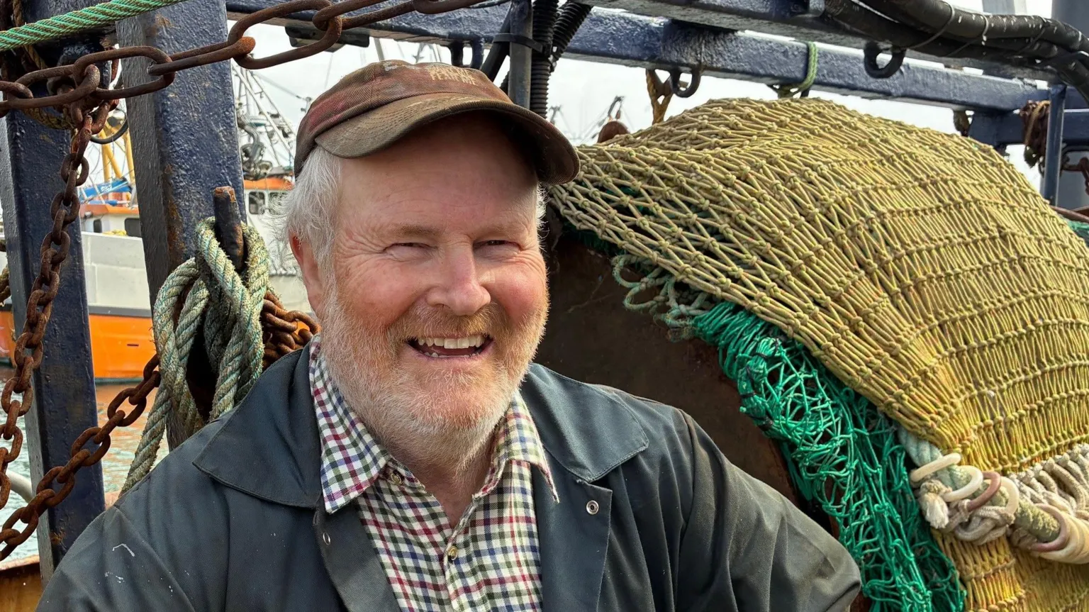 Tom McClure sat on his vessel smiling at the camera. Behind him is a yellow and green fishing net. He is wearing a brown cap, checked shirt and black jacket.