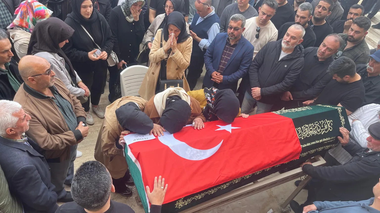 A solemn-looking crowd gathers around a coffin, which has the Turkish flag draped over it, while three women wearing head coverings rest their heads on the coffin.