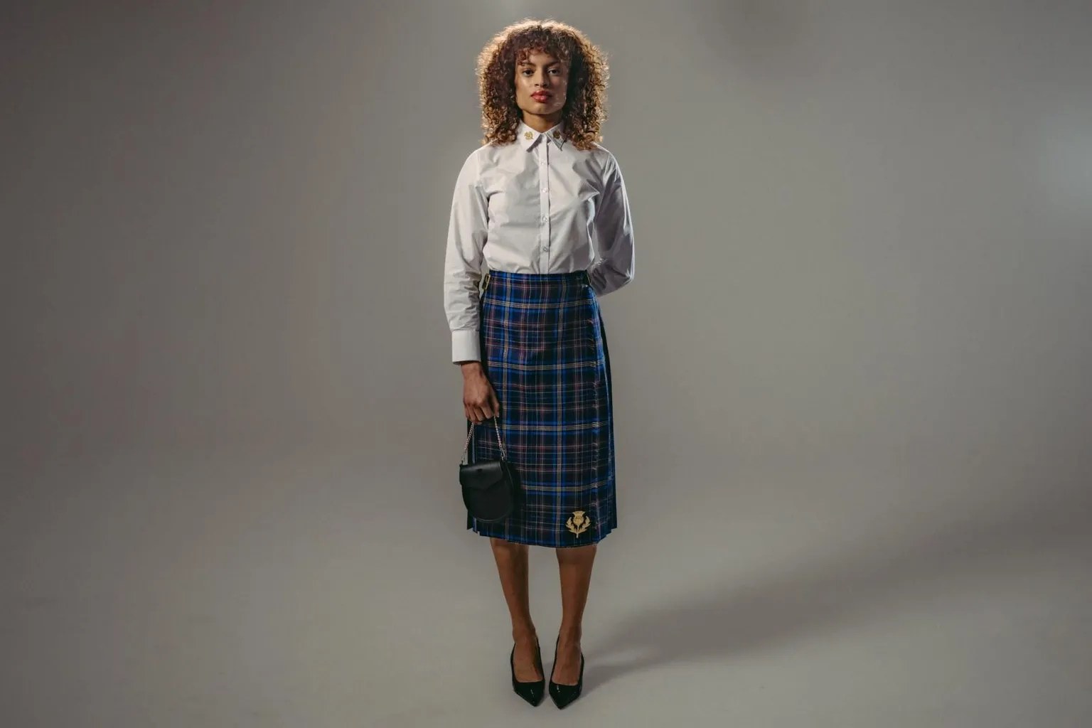 Team Scotland A female model who is wearing a long, blue kilt and a white shirt, with black shoes and a small, black bag.