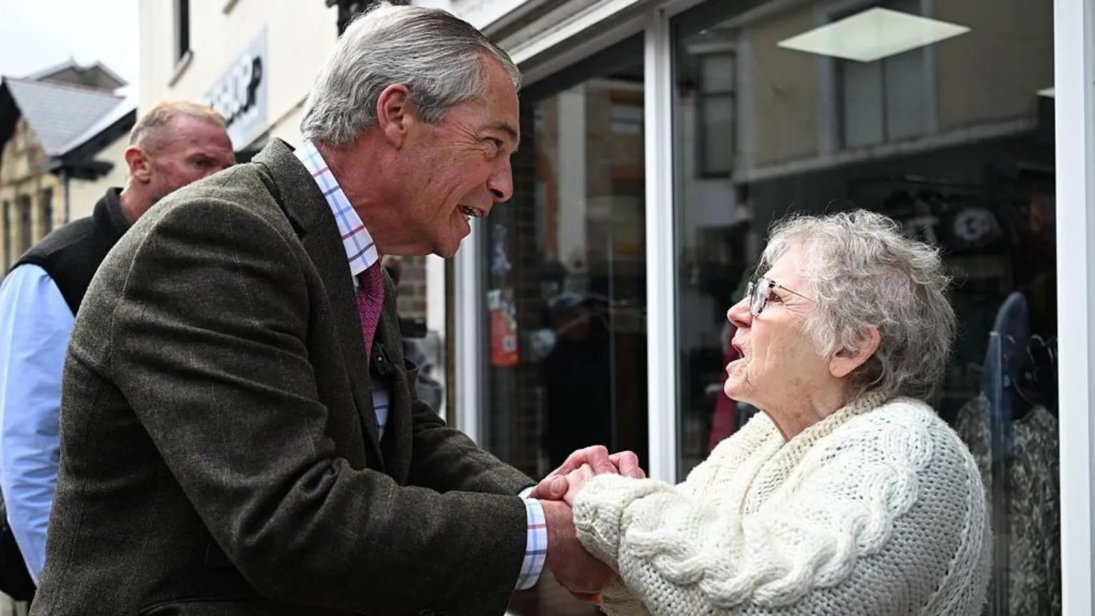 Matthew Horwood/ Nigel Farage talking to an older woman in a white cardigan outside a shop.
