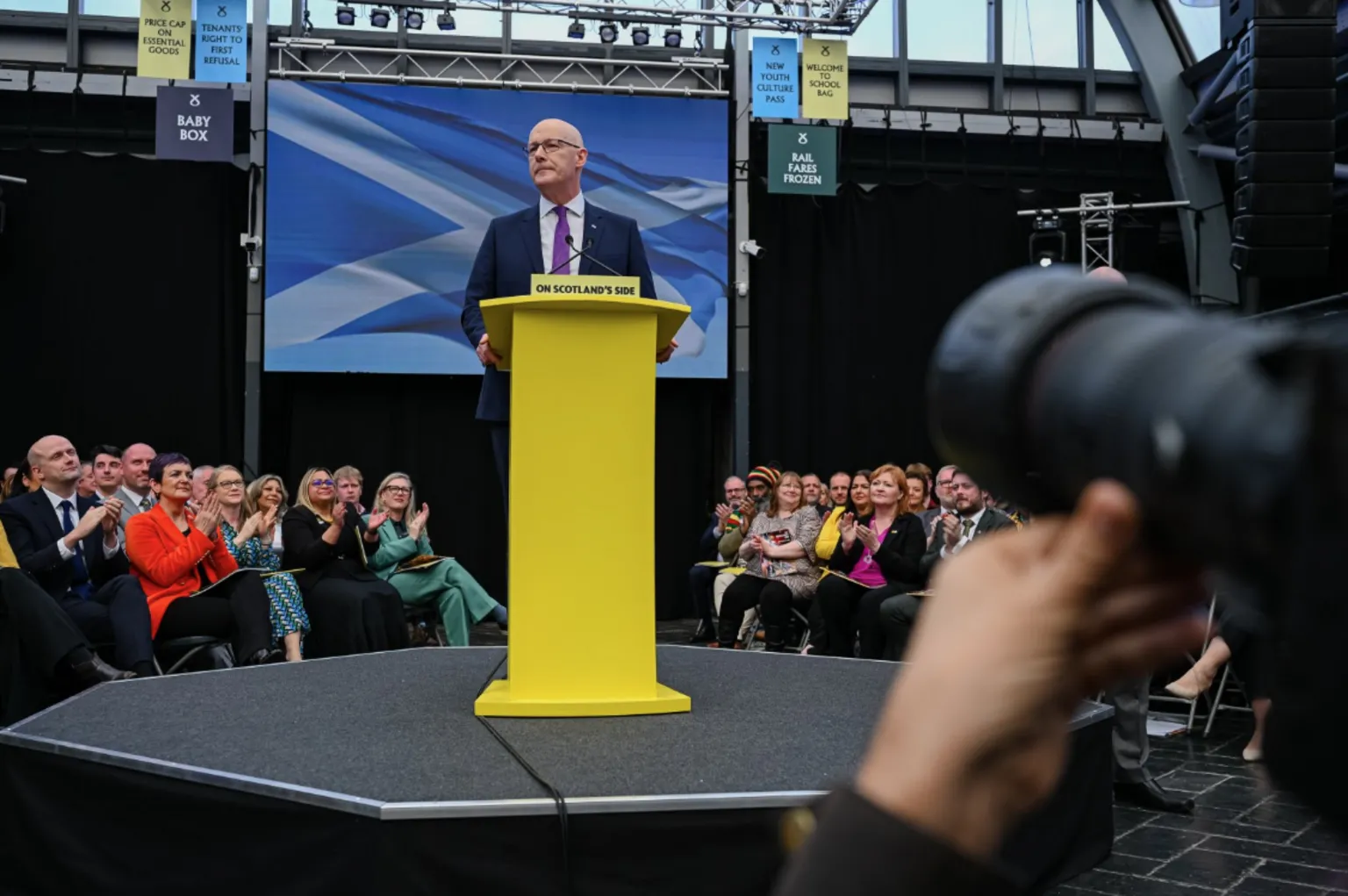  John Swinney standing at a bright yellow podium on a small stage inside an indoor venue, speaking to an audience seated around the stage, with a large blue-and-white backdrop behind the podium and a camera lens visible in the foreground.”