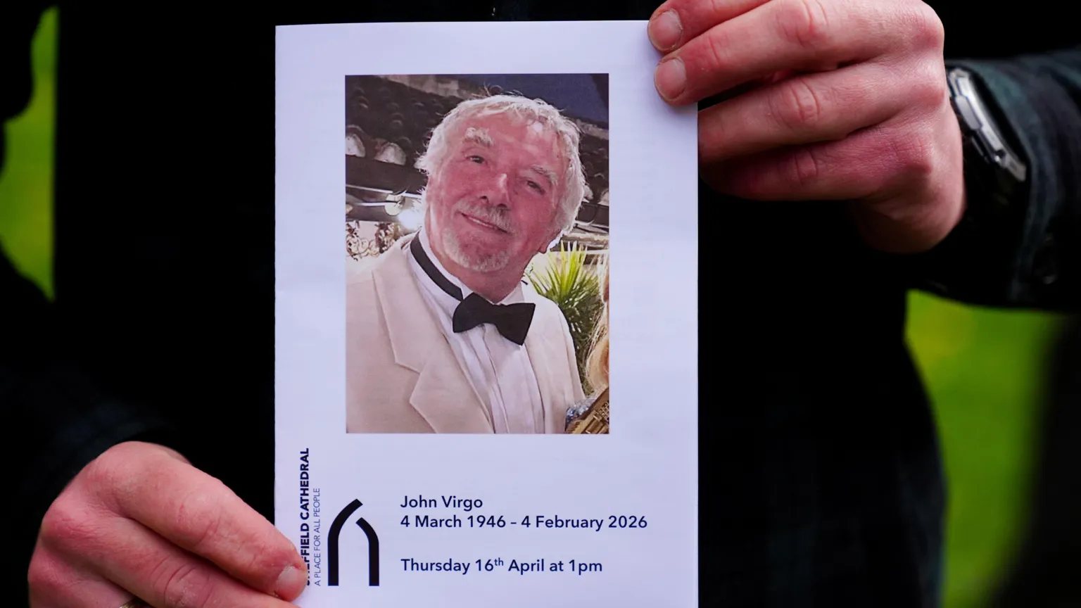 A pamphlet advertising the memorial service for John Virgo is held up by a pair of hands.