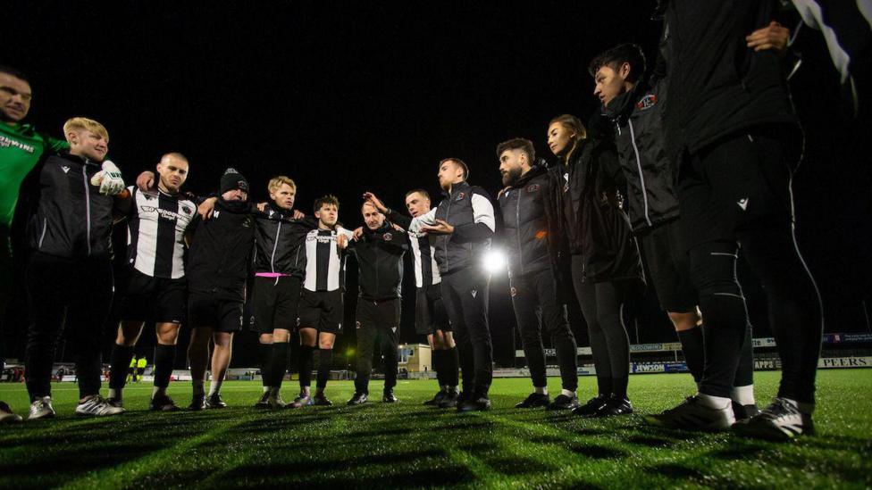 Llandudno players in a team huddle on a floodlit pitch