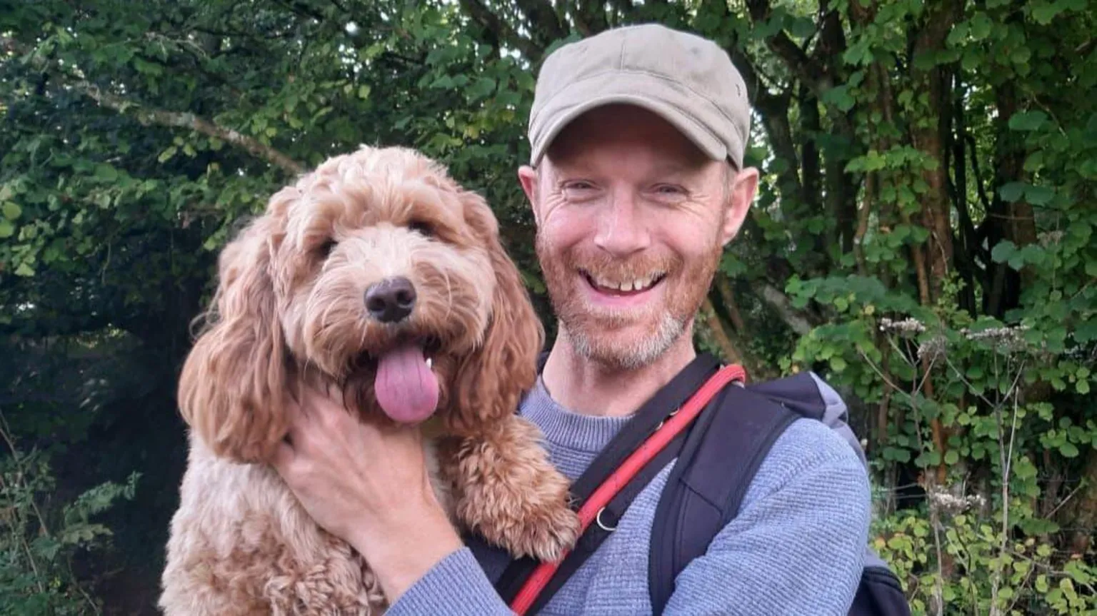 Lee Mason Lee Mason posing holding a dog in his arms whilst stood in a park. He is wearing a purple jumper and a beige cap.