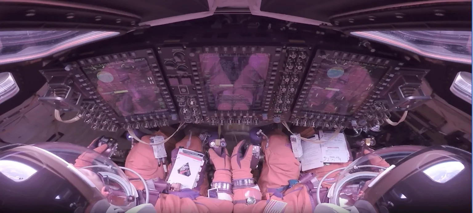 NASA This image looks forward over the shoulders of two Artemis II astronauts strapped into their seats inside the Orion crew module. They wear bulky orange pressure suits and clear helmet bubbles, facing a wall of tall, glowing display screens and dense rows of switches and knobs. Their gloved hands rest near hand controllers and a checklist. Around the edges of the frame, curved windows, cables and equipment make the cabin look compact and highly technical