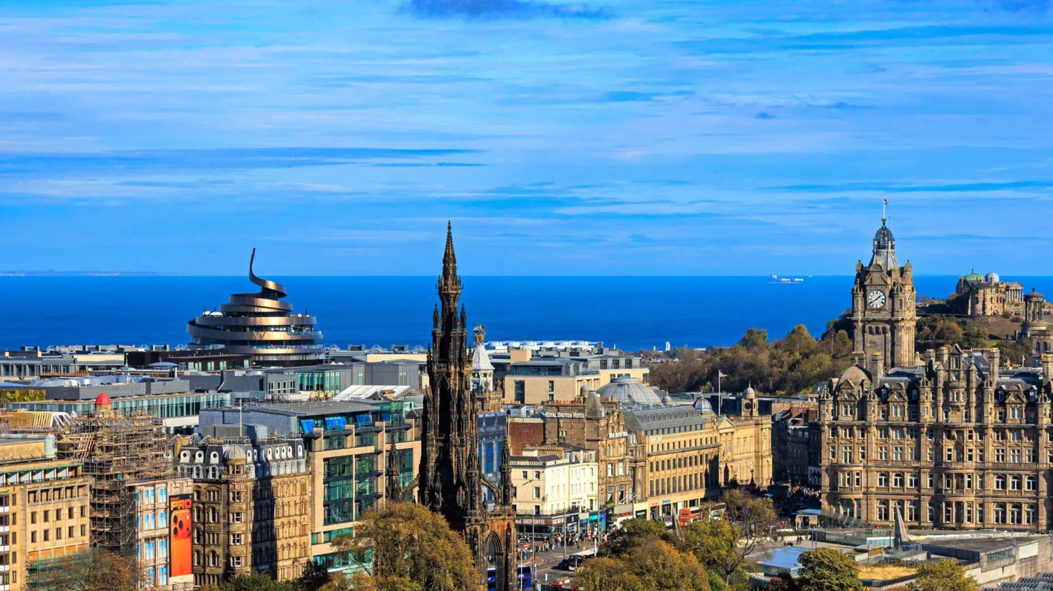  A picture of the central Edinburgh skyline, with buildings including the Scotsman Hotel and Scott Monument standing out against a blue sky over the blue water of the Forth