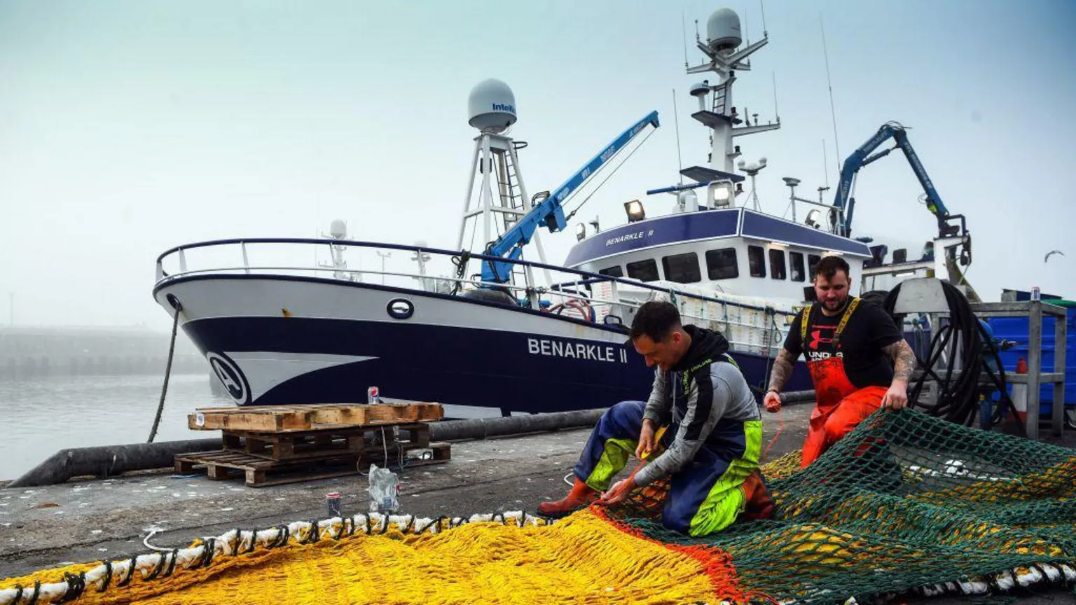  A picture of a fishing boat at Peterhead harbour, with two men in high-visibility wading trousers mending nets in the foreground