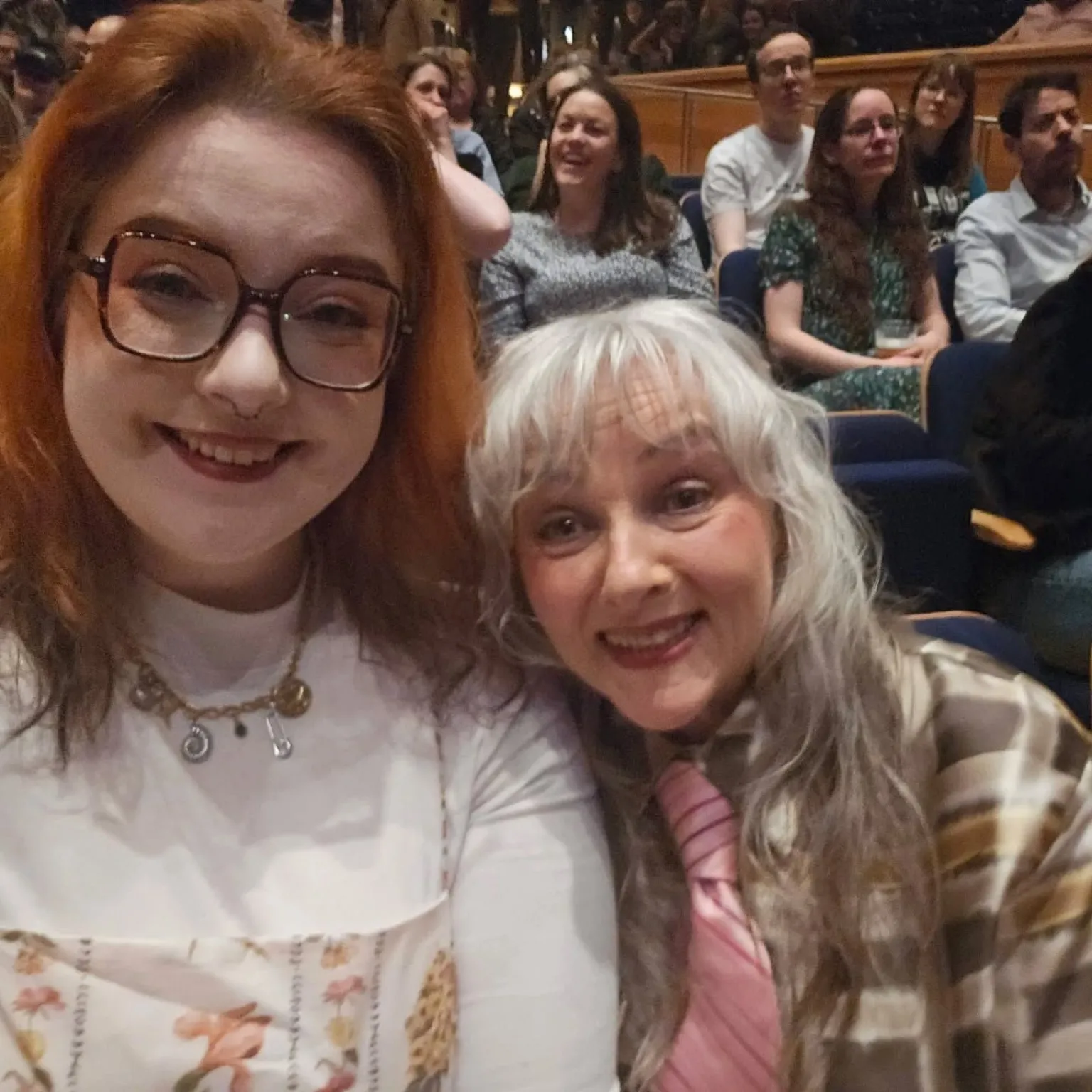 Anita West Two women sitting in the seats at a concert - one woman has long red hair and a white top on. The other has blonde/silver hair and a patterned top in shades of beige and brown