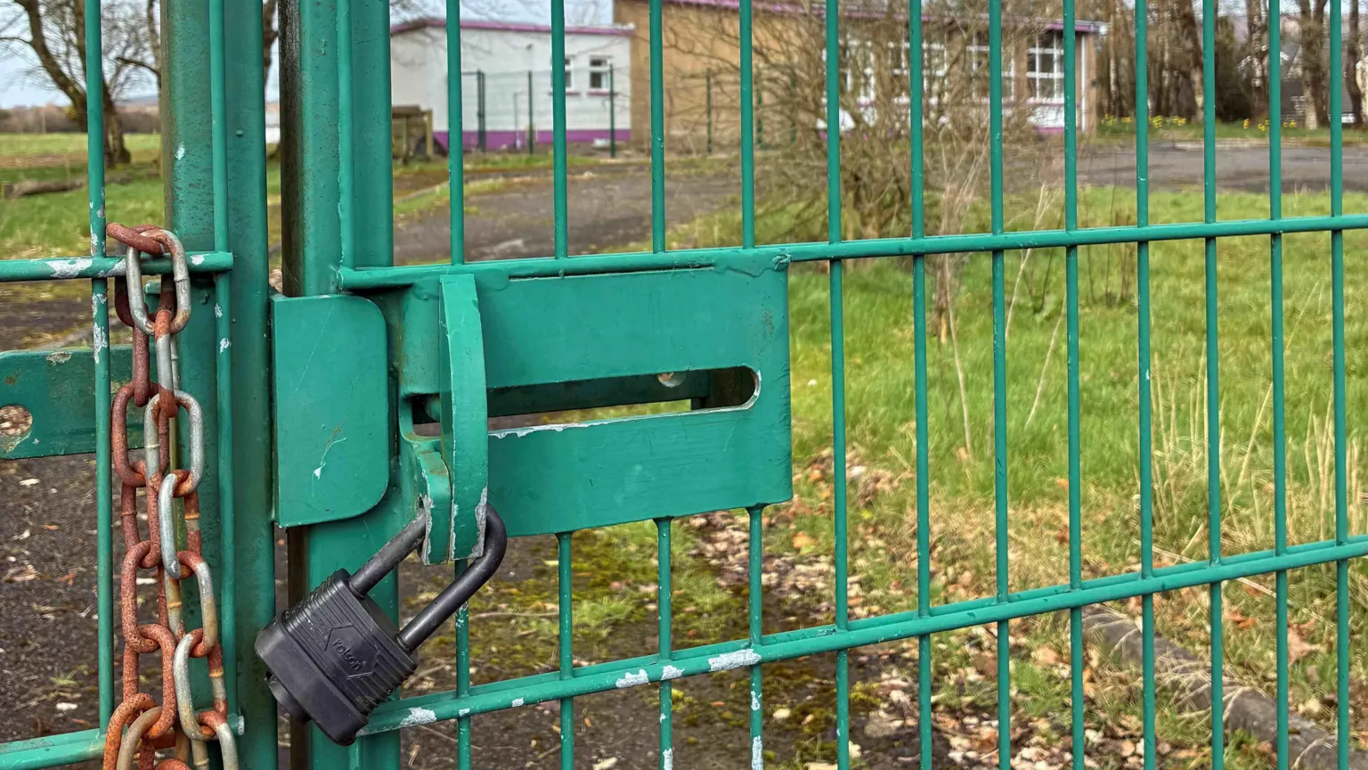 A green gate with a locked black padlock. Behind the gate is a small building with grass and trees around it. 