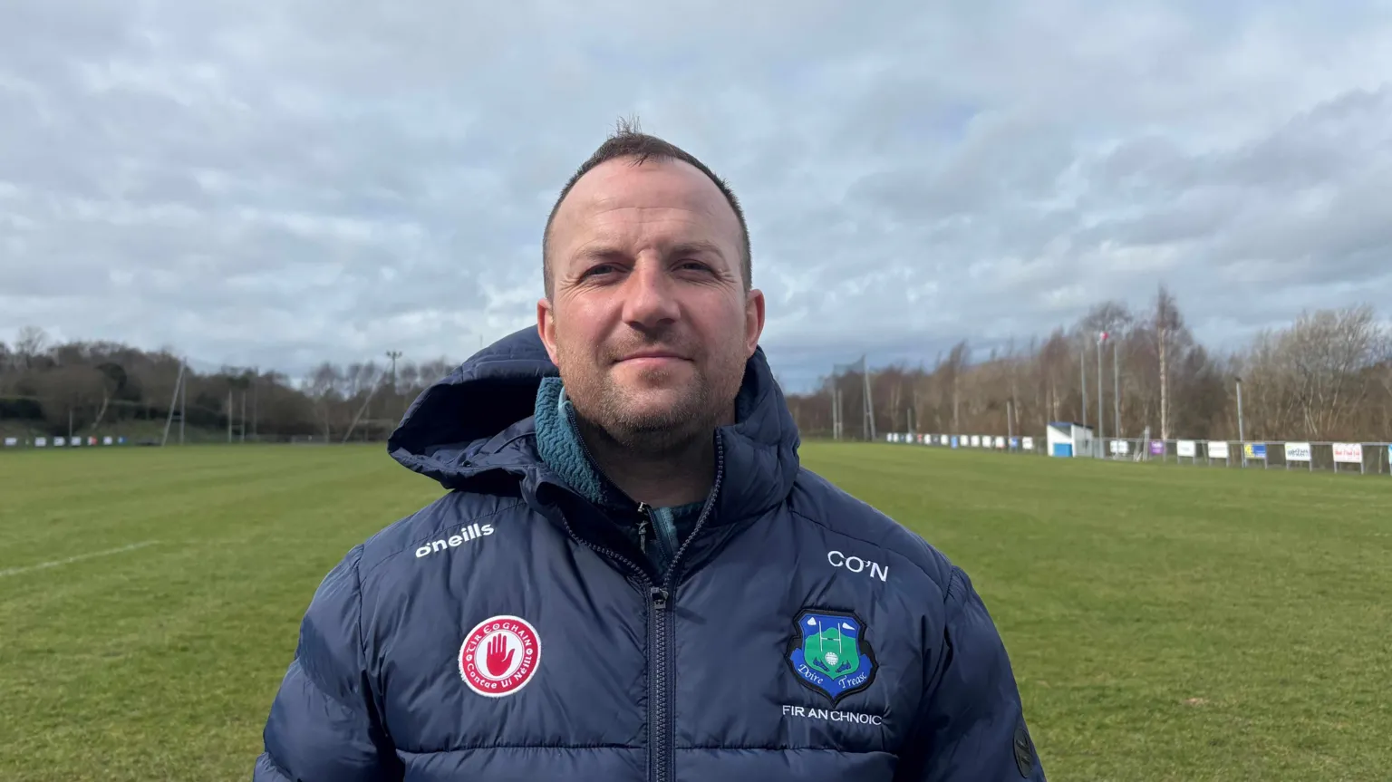 A man wearing a dark jacket with several sport badges on it. He has short brown hair and is looking at the camera. He is standing on a pitch. There are trees in the background. 