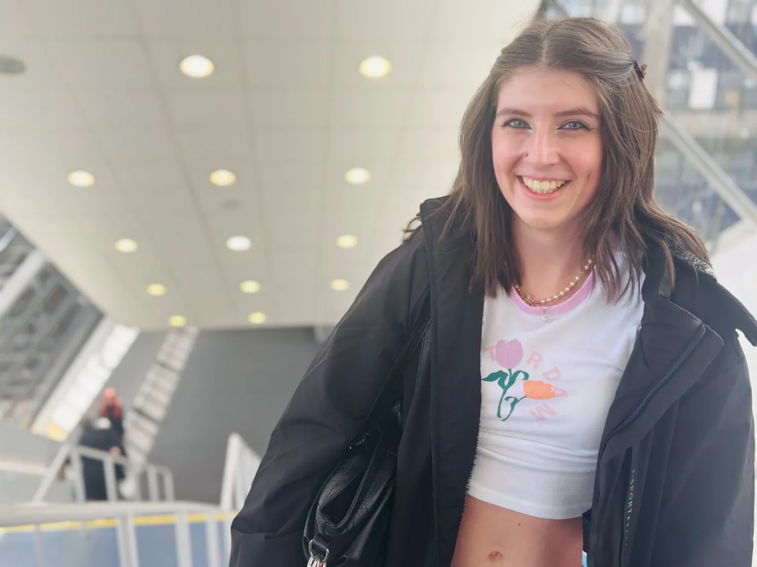 Paige Jackson, a teenage girl, smiling at the camera at the top of stairs, she had dark hair and is wearing a light cropped top and a dark jacket.