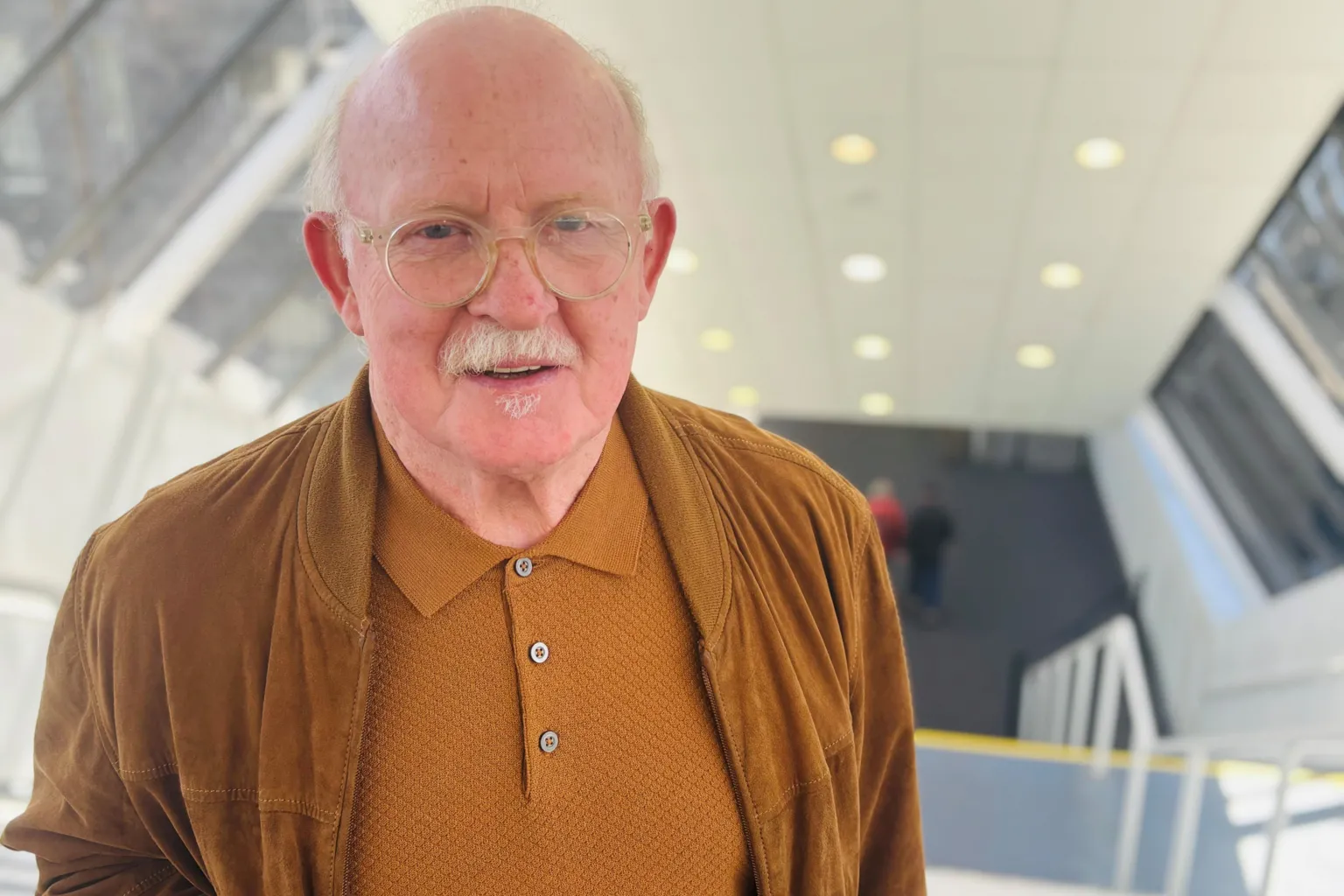 Kenneth Wilkie, a man in a brown top and jacket, smiling at the camera, at the top of stairs