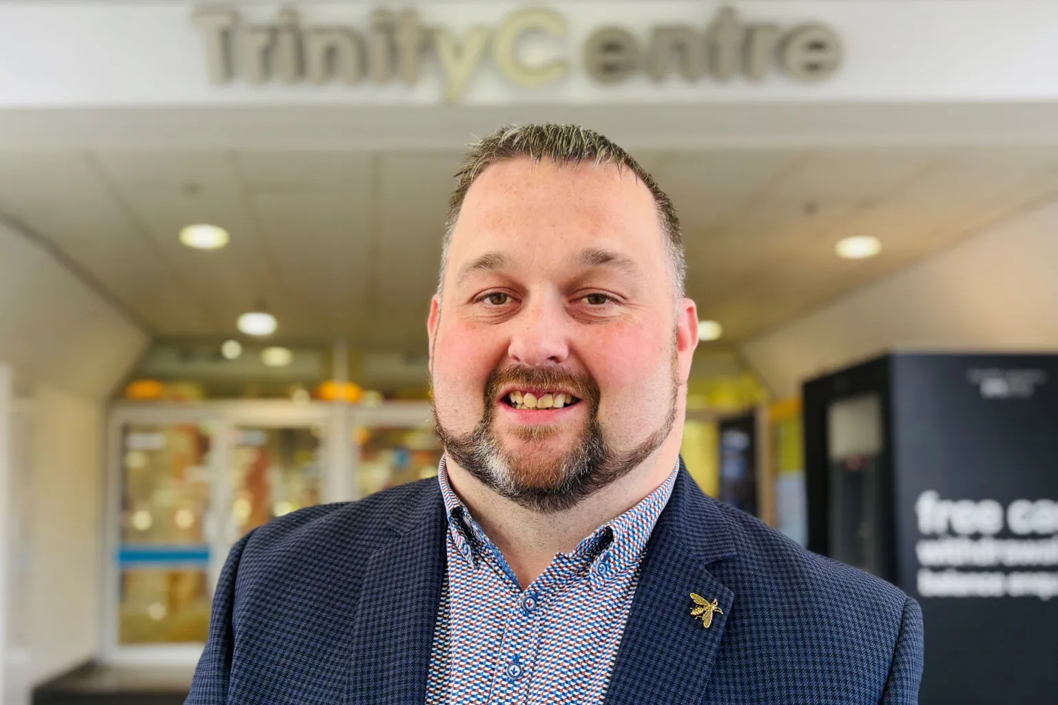 Kenny Bruce smiling at the camera, he is wearing a blue suit jacket and a patterned shirt, the words Trinity Centre are behind him on a sign.