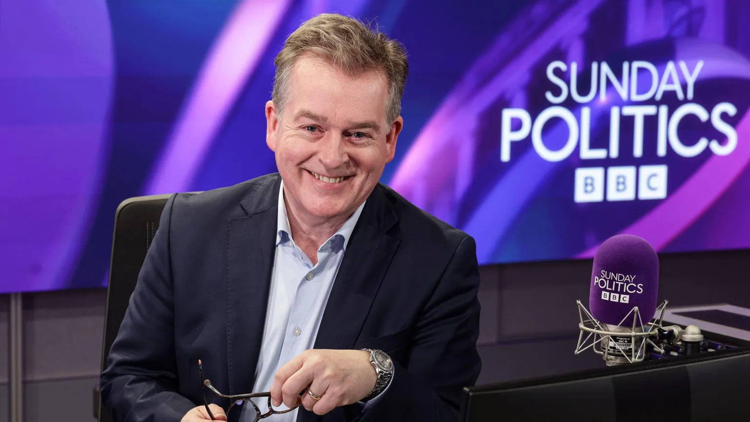 Mark Carruthers is holding his glasses and smiling. He's sitting on a computer chair at a desk in a studio. Sunday Politics BBC is displayed behind him on a screen and next to him on a mic. He's wearing a navy suit with a light blue shirt and a silver watch. 