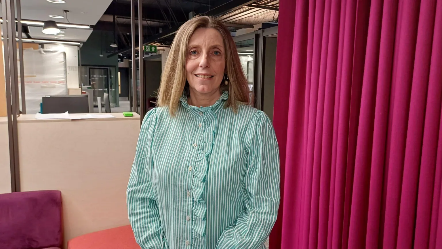 Gwyneth Jones is standing in an office. She is wearing a green and white stripped top and hoop earrings with a pendant on them. There is a pink curtain behind her. 