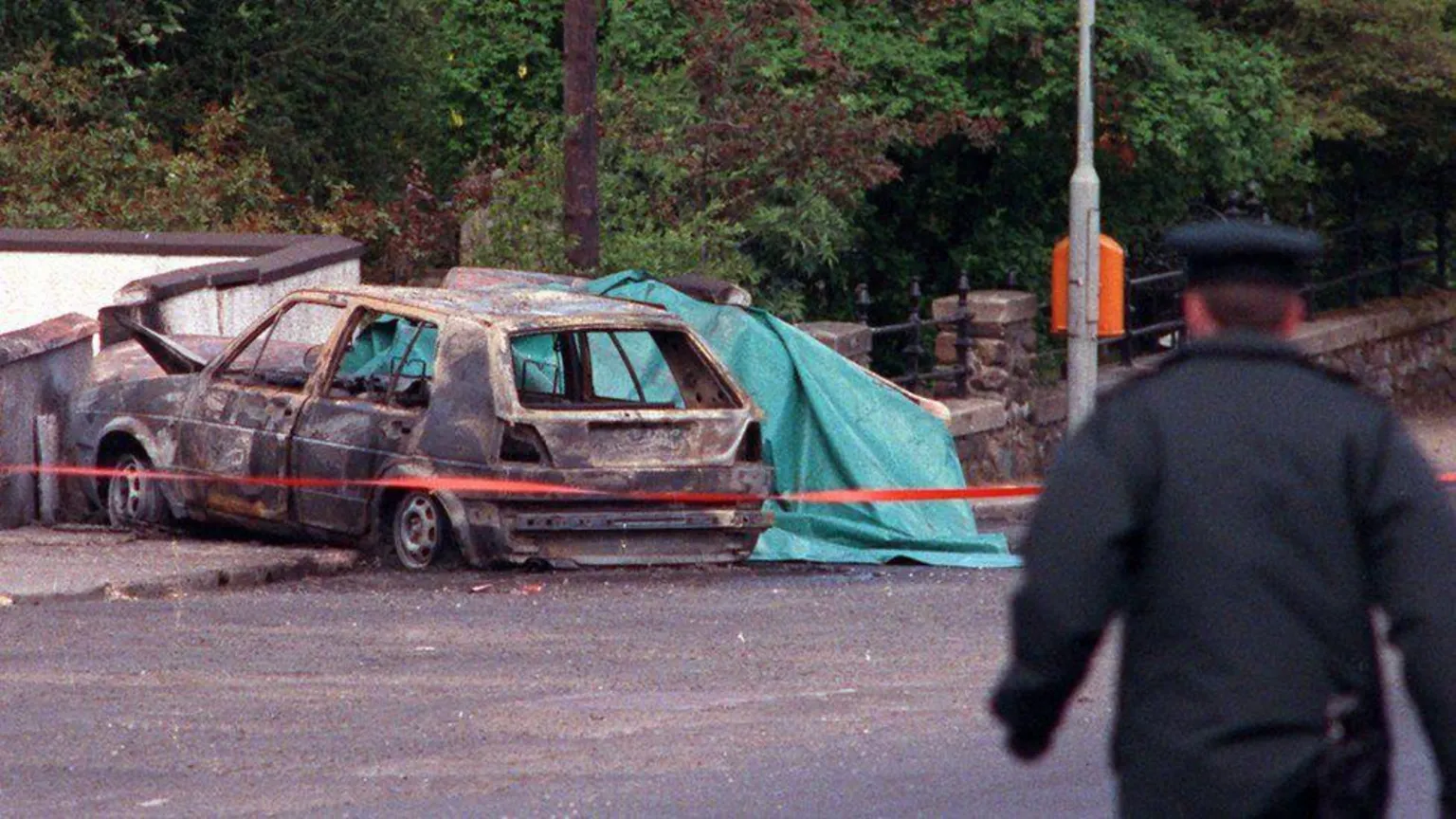 Pacemaker A car burned out, there is red tape blocking it off and a police officer with his back to the scene in a heavy coat and peaked cap. The car sits beside a stone wall.