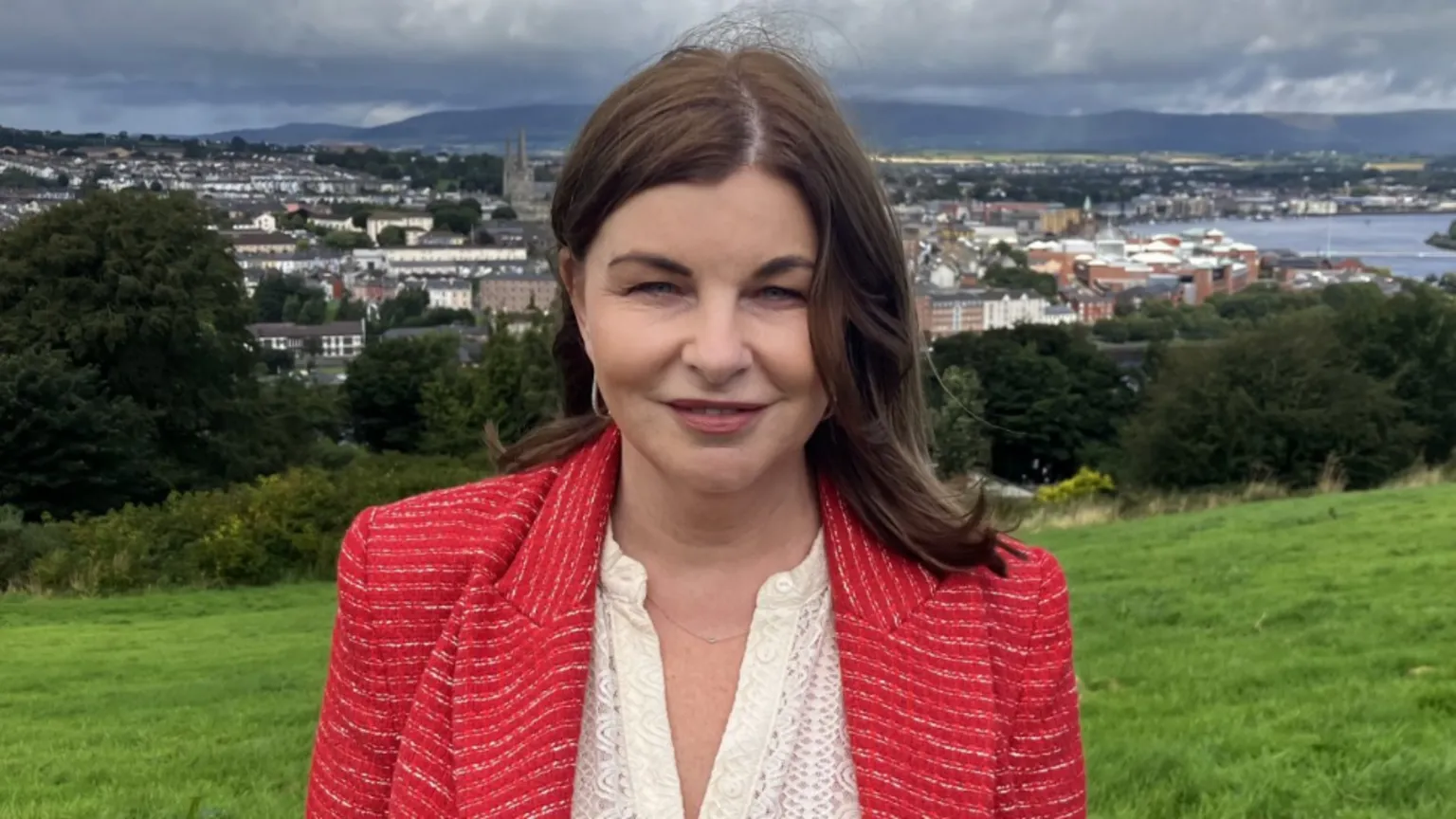 Shows Sinead McLaughlin in a red blazer with a cream open neck top standing in a field with trees in the background and a river and buildings beyond that.