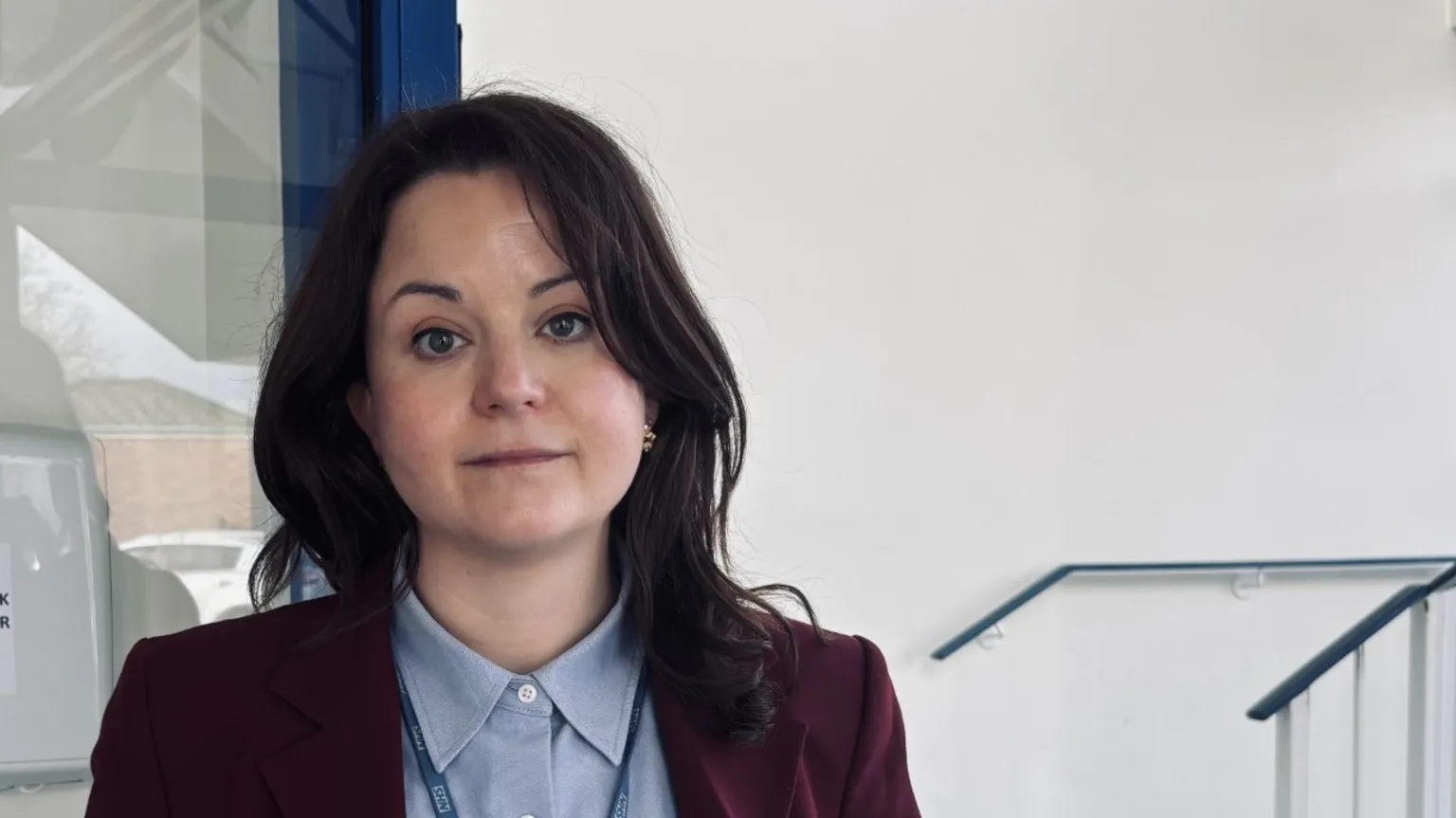 A woman with wavy dark hair to her shoulders. She is wearing a pale blue button-up shirt and maroon blazer. She has an NHS lanyard around her neck and is looking at the camera with a neutral expression. She is stood in front of a plain white wall. 