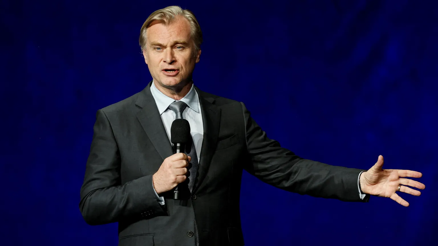  Christopher Nolan in a suit speaking on stage into a hand-held microphone with the other arm outstretched