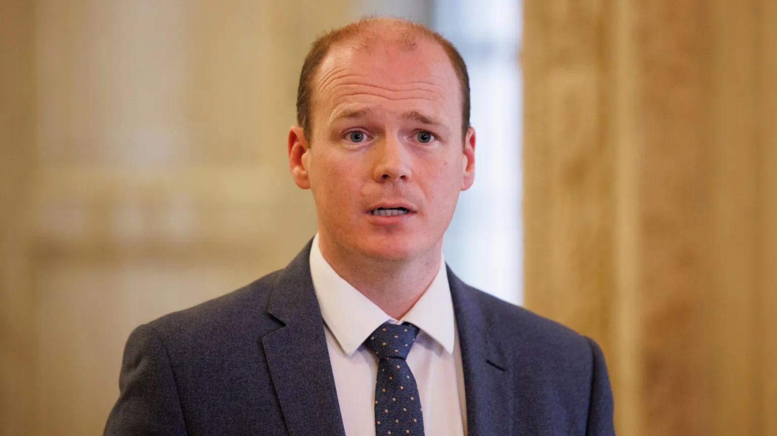  Gordon Lyons is balding with reddish/brown hair at the sides and is wearing a dark suit and tie with a white shirt