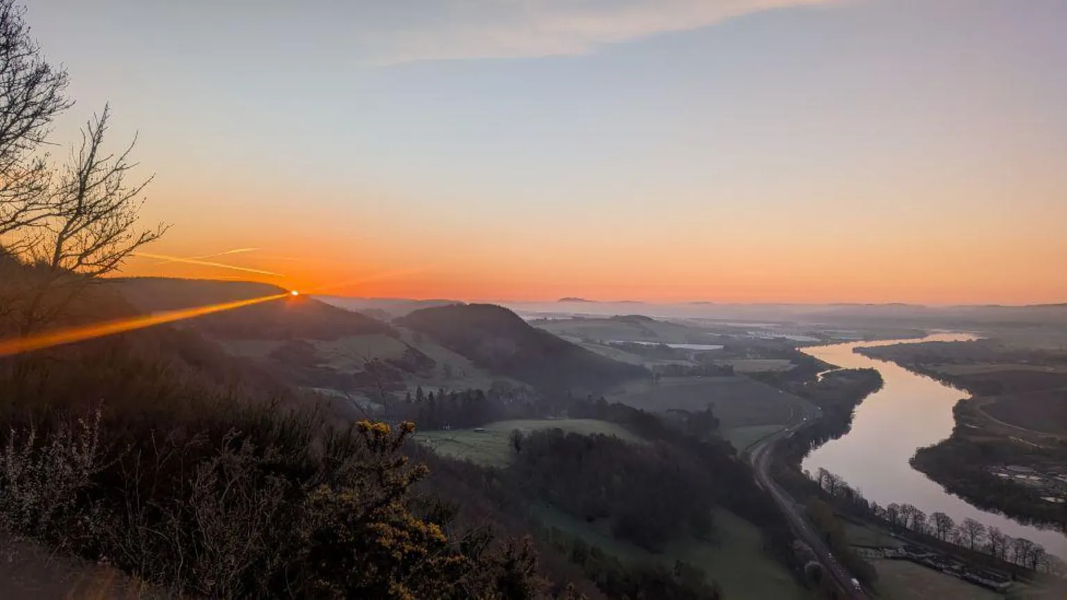 Rich Garside Organe sunrise glow appearing over hilltops in the distance, with a winding river and field and trees also in the shot. 