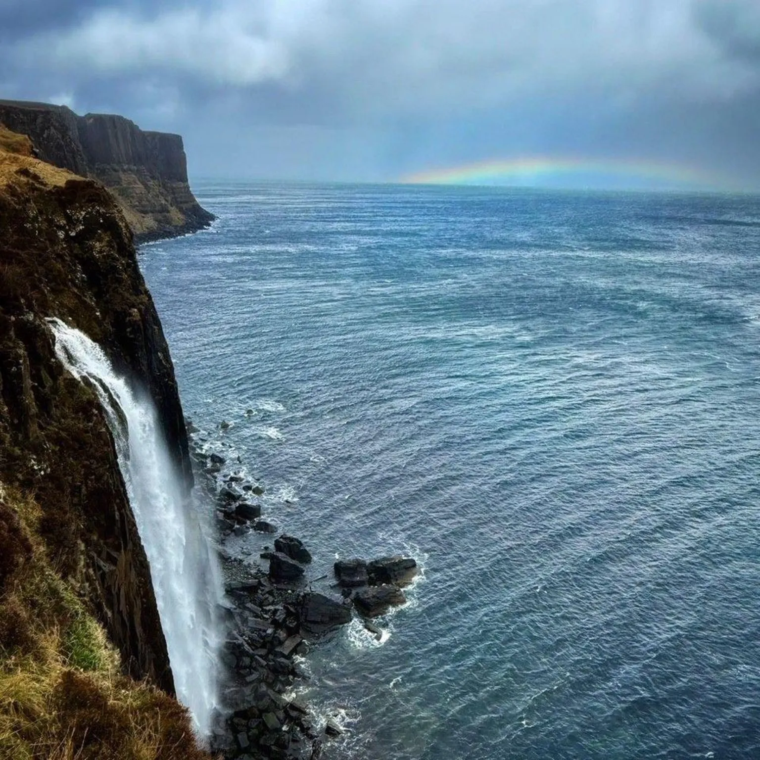 Peter Xu A distant rainbow over the sea, with a waterfall going into the water in the foreground.