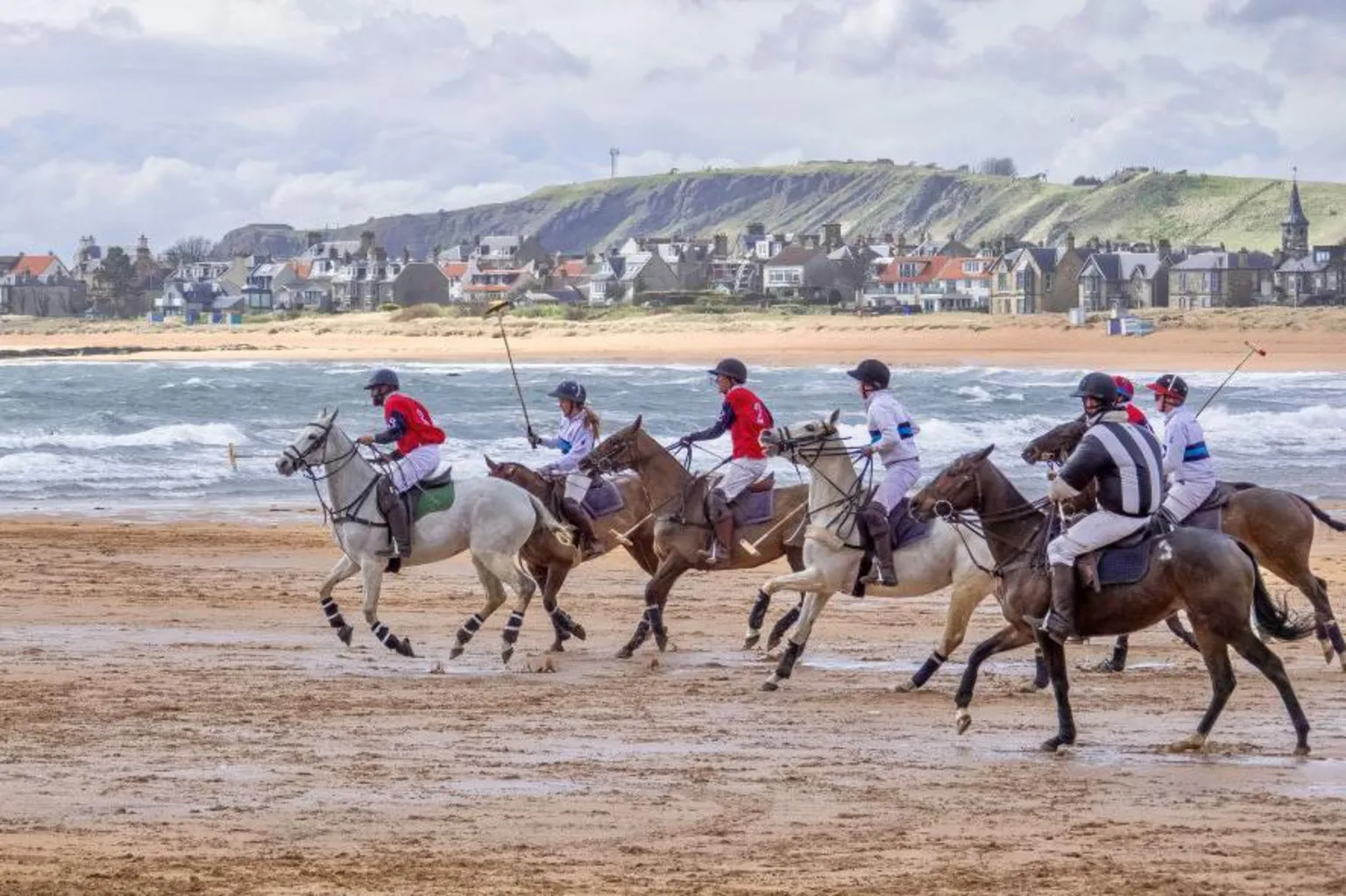 Bill Bruce Seven people playing polo on horseback on a beach, sea in the distance, and houses in the distance.