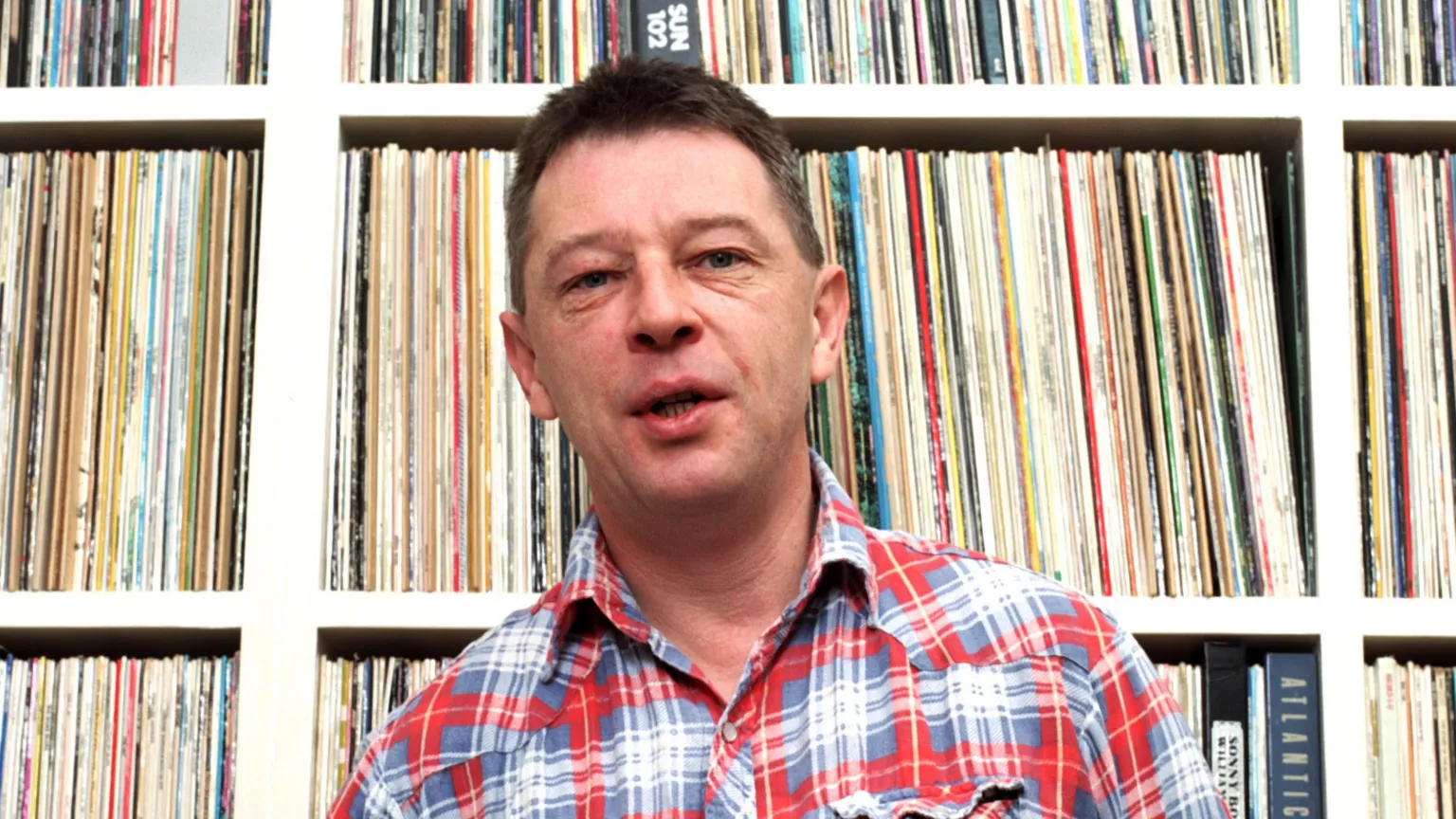  Andy Kershaw in a checkered shirt looking at the camera while in front of his record collection
