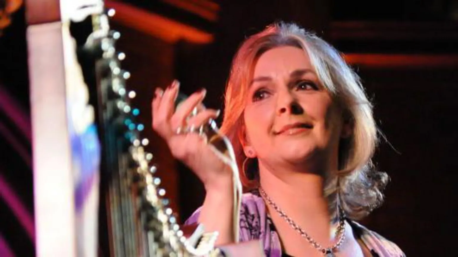  Moya Brennan is standing beside a harp. She is wearing a silver necklace and has long blonde hair. Her right hand is raised.
