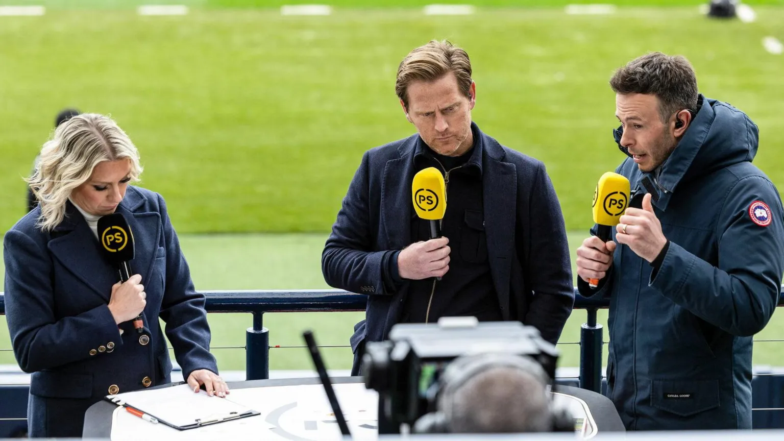 SNS Three pundits, a woman and two men, stand facing a TV camera holding yellow and black microphones which read 