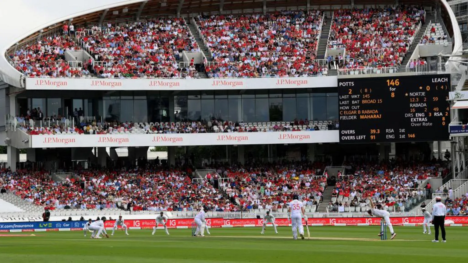  A wide view of a cricket match in progress at Lord’s Cricket Ground, showing players on the pitch and a packed grandstand with spectators, sponsor signage, and a large electronic scoreboard in the background.