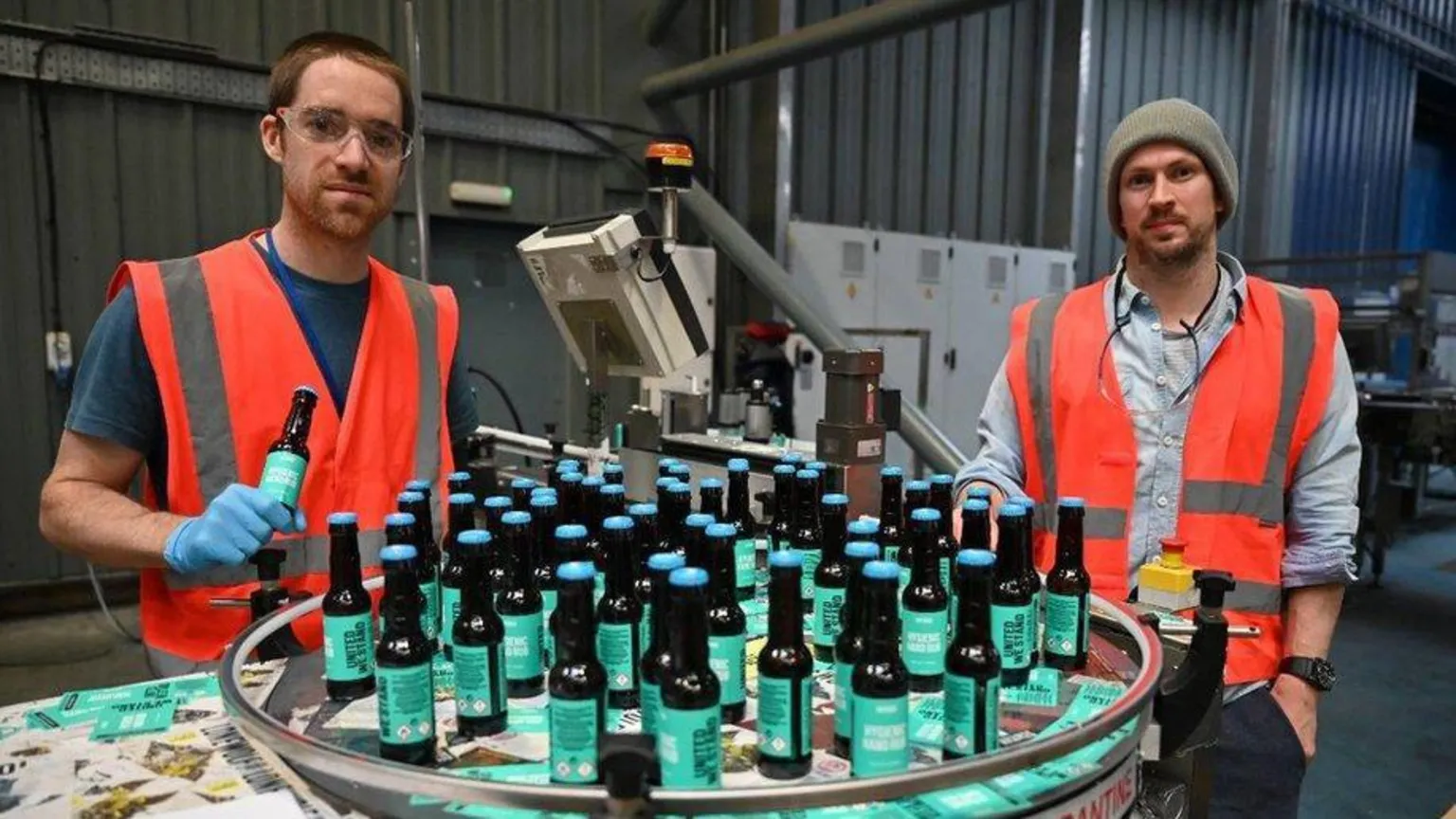  Dickie and Watt wearing high‑visibility vests stand beside a circular bottling table filled with BrewDog beer bottles inside an industrial brewery, with production equipment visible in the background.