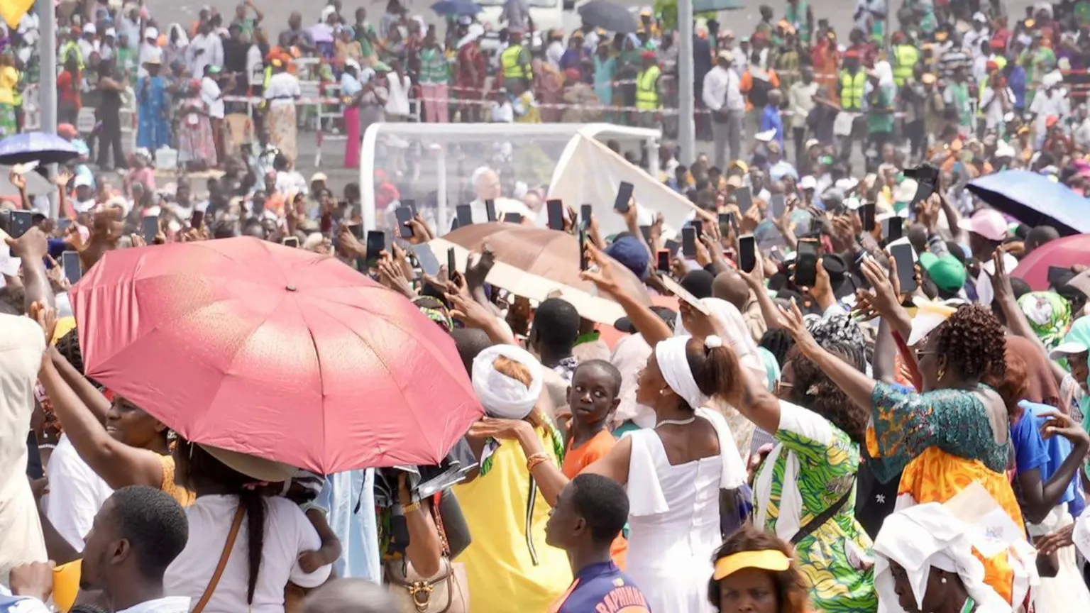 Michel Mvondo/BBC Crowds with their back to the camera facing the popemobile. There are some umbrellas in the crowd to shade against the sun, most notably a red one on the left. Many people in the crowd have their phones up in the air to take photos or videos.