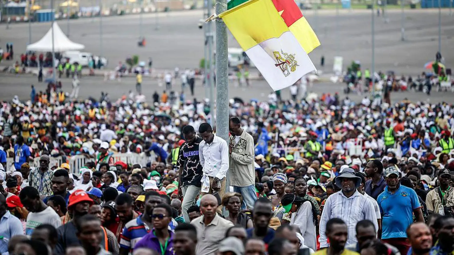 AFP via Crowd of people with a pole in the centre with three men stood against it. the pole has a Vatican flag and a Cameroon flag attached. They all look to be listening