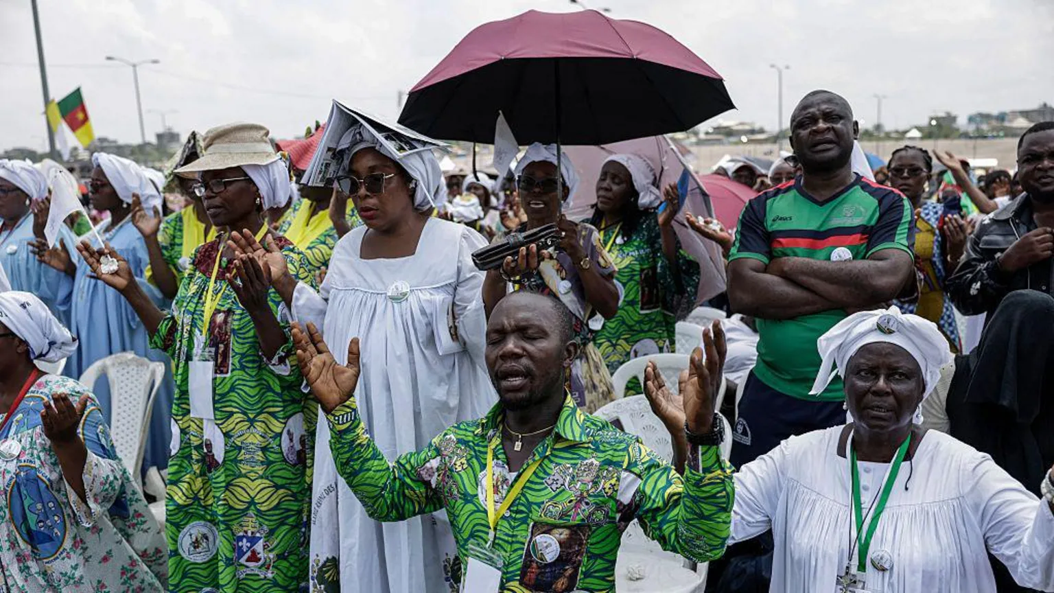 AFP via A man in a vibrant green shirt with his eyes closed and hands raised in prayer. He is kneeling as is the woman on his left. Behind them are dozens of people standing, including one with a cap on, one with an umbrella up, and a woman with an open book over her head, all to shield from the sun.