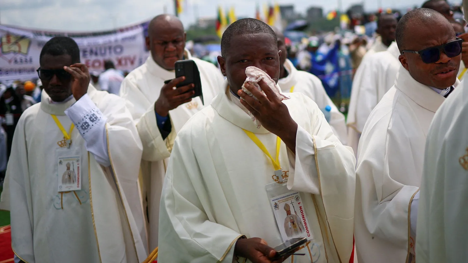  Four clergymen, in front of a crowd. The one in the centre is wiping sweat from his face with a hankerchief. 