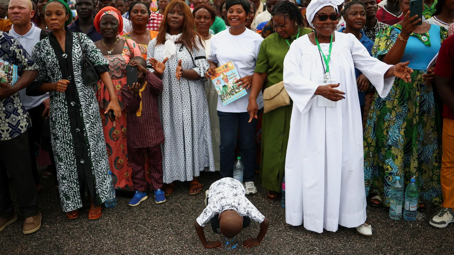  A crowd of happy people standing. In the front centre is a little boy who is prostrating on the ground