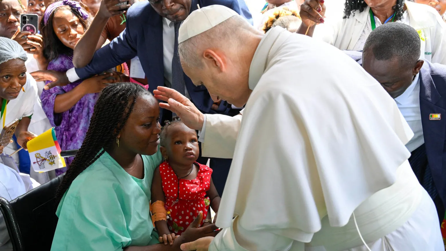  The Pope is blessing a woman in green, who is holding a toddler girl on her lap. There is a crowd of people around them