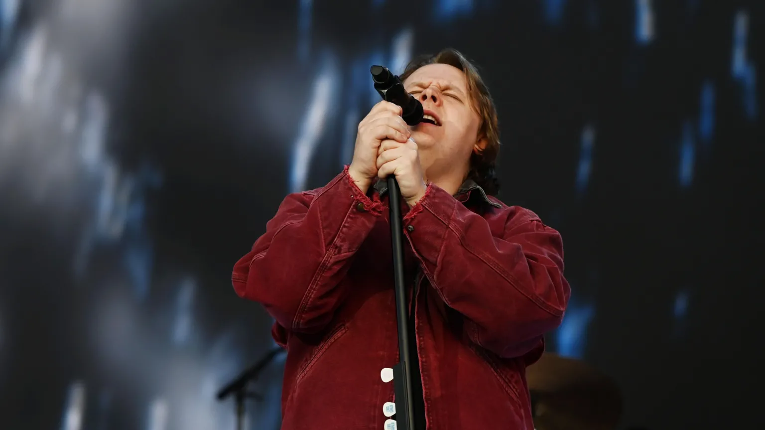 Lewis Capaldi singing onstage, wearing a burgundy jacket. He has short brown hair and is clutching the microphone stand intensely.