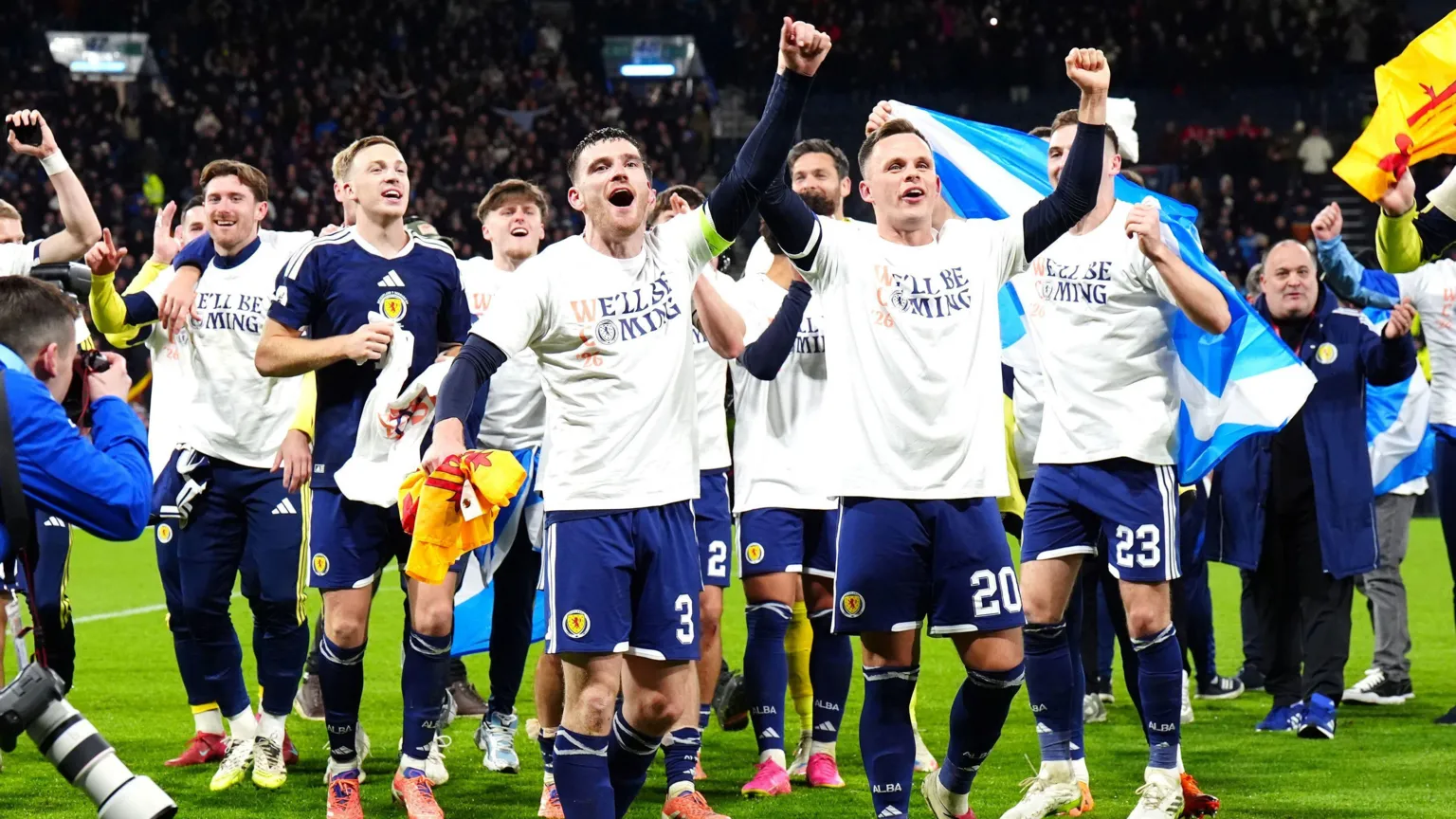  Scotland players celebrate after reaching the World Cup. Several have their arms raised joyfully while others salute the fans. Most of the team are wearing T-shirts saying We'll Be Coming.