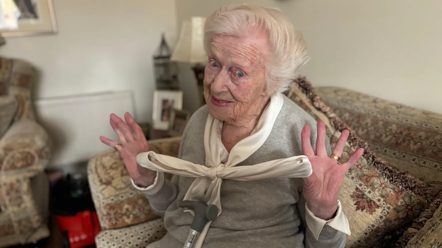 An older woman sitting on her couch holds up the bow in her shirt. She has white hair and blue eyes, and a crutch resting against her as she sits.