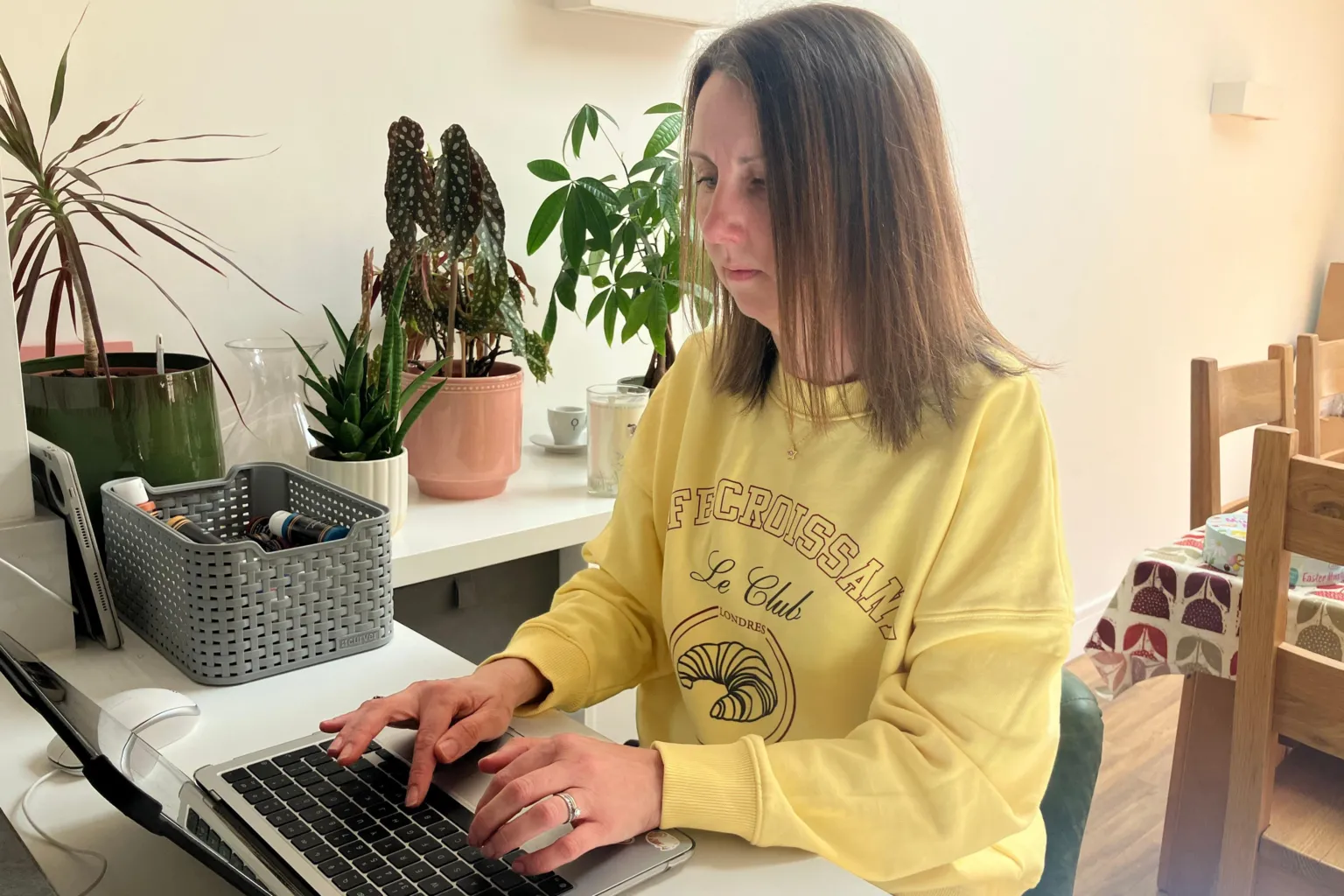 Leanne Rinne / BBC A woman with medium-length brown hair wearing a bright yellow jumper typing on her laptop, she is sat down on a green chair. 