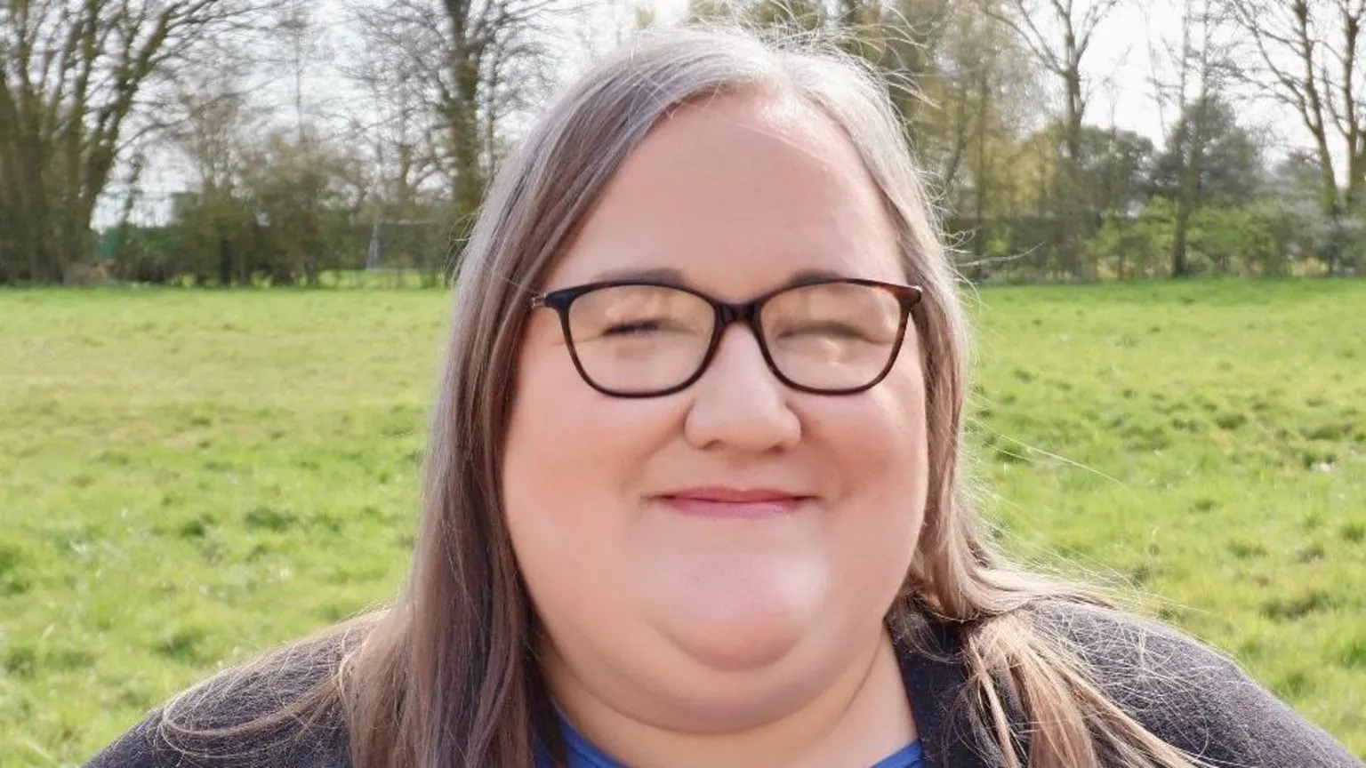 Becky Alderton Becky Alderton stands in a grass field and smiles at the camera. She has long brown hair and wears glasses.