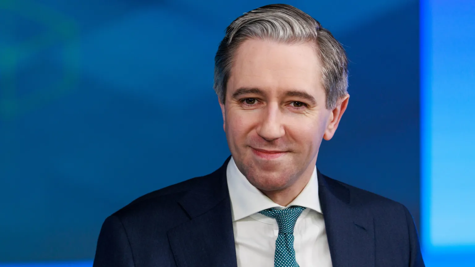  Simon Harris is smiling. He's wearing a navy suit with a white shirt, and a blue tie decorated with white spots. His hair is black and white and brushed to the side. The background is blue and out of focus. 