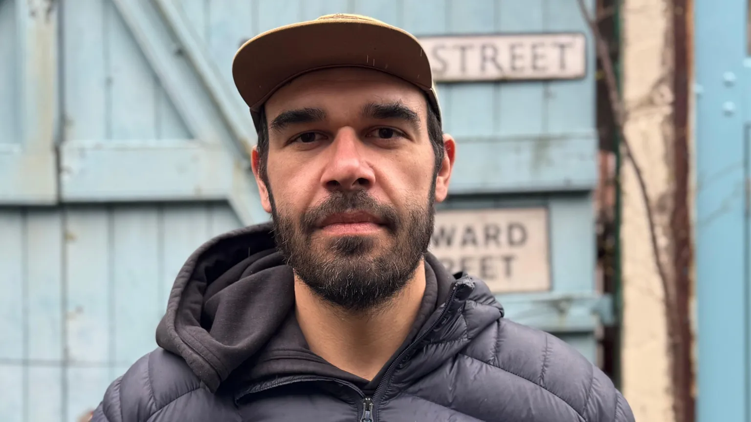 Daniel is standing outside in front of a blue fence. He is wearing a black cap and coat. He has a black beard. 