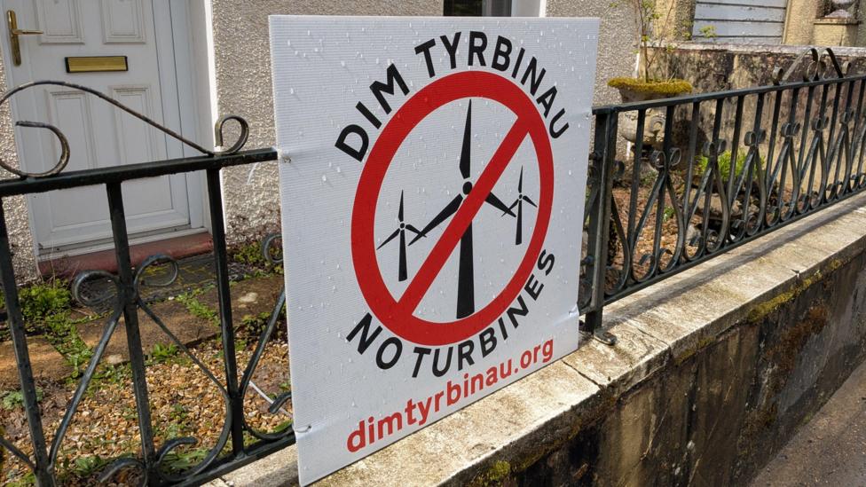 Poster ar glwyd o flaen tŷ ym mhentref Rhydcymerau sy'n dweud 'Dim Tyrbinau'. Mae'r poster yn dangos tyrbinau ac arwydd coch drostyn nhw, yn cefnogi eu gwahardd.