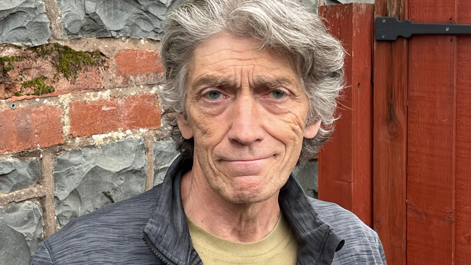 Ian Young is standing in front of a wall made of bricks and stone, as well as a large red wooden gate. He has grey hair and is smiling at the camera.